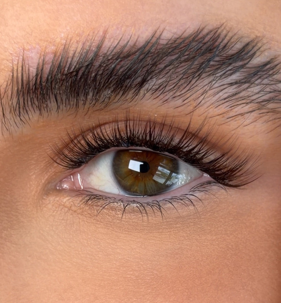 Close-up of a person's eye with a dark, well-defined eyebrow and long eyelashes, with a hand holding a white cosmetic tool near the eyebrow.