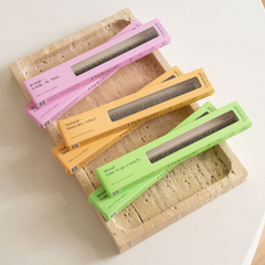 Four boxed eyelash extensions in pastel pink, orange, yellow, and green, arranged diagonally on a beige travertine tray against a white background.